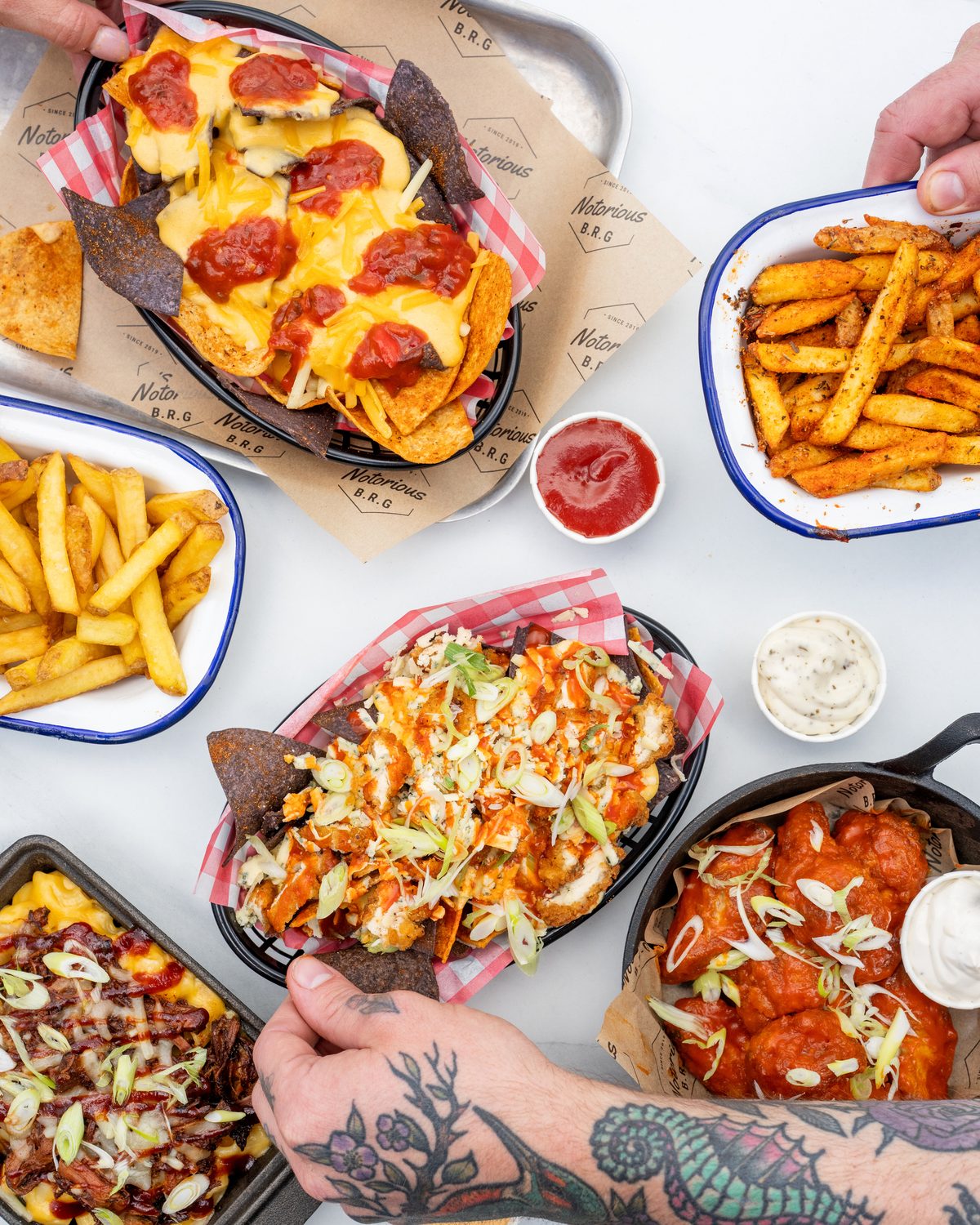 Seen from above: loaded nachos, fries, wings and mac spread across a table, with hands reaching in to share